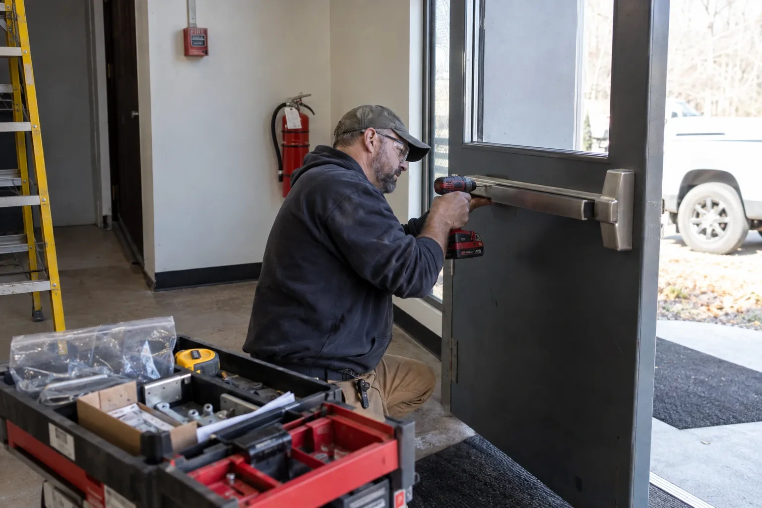 Technician in safety glasses kneeling at a gray commercial door, drilling a steel push bar into place beside an open parts case.